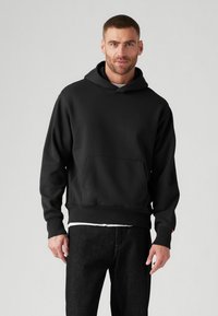 Sudadera negra de tela suave, con un bolsillo frontal, cuello ajustable y puños acanalados. Se lleva con pantalones de mezclilla oscuros.