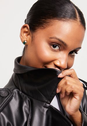 Chaqueta de cuero negra con un cuello alto, textura suave y detalle de botones. La modelo lleva un pendiente dorado con una gema verde, parcialmente cubierto por el cuello.