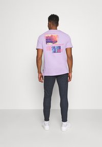 Ljuslila t-shirt med grafiska tryck i röd och blå på ryggen, tillsammans med mörka åtsittande byxor och vita sneakers.