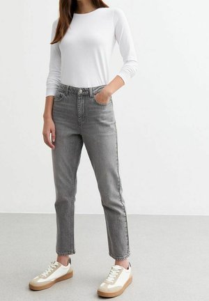 Kvinde iført hvid langærmet skjorte, højttaljede grå jeans og hvide sneakers med beige accenter, stående mod en ensfarvet hvid baggrund.