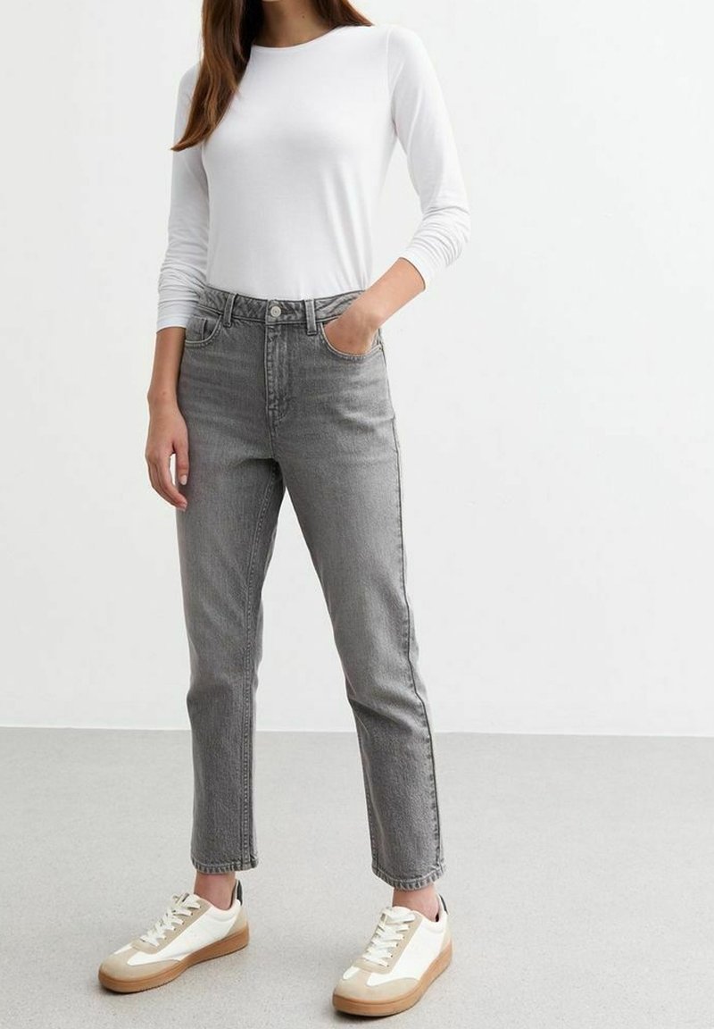 Femme portant une chemise blanche à manches longues, un jean gris taille haute et des baskets blanches avec des accents beige, debout devant un fond blanc uni.