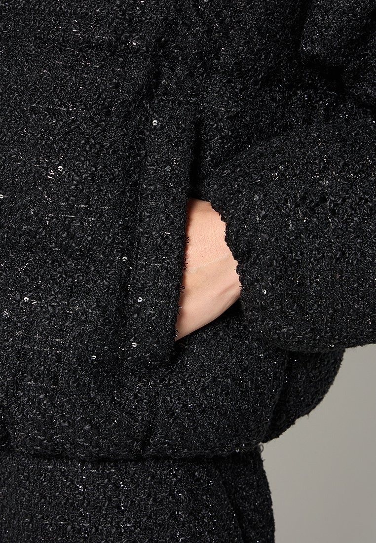Schwarzer, strukturierter Stoff mit einem glitzernden Finish; verfügt über eine Handtasche und einen lockeren Schnitt, der das weiche, gemütliche Erscheinungsbild betont.