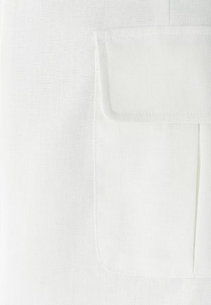 Bela tkanina z teksturiranim tkanjem, s patno žepom na levi strani in gubanjem pod žepom.
