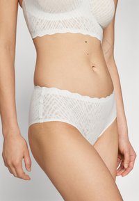 Fehér csipkés bralette és hozzá illő hipster alsónemű szett hullámos széllel. Texturált, geometrikus mintázattal rendelkezik, ami vizuális érdekességet ad.