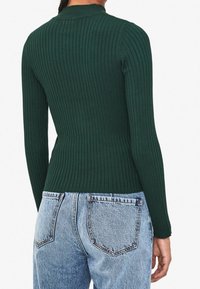 Dunkelgrüner gerippter Pullover mit taillierter Passform und langen Ärmeln, von hinten betrachtet, kombiniert mit hellblauen Denim-Jeans.