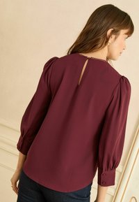 Bordinės spalvos palaidinė su pūstomis rankovėmis, raktelio formos atidarymu nugaroje ir rankogaliais su sagutėmis. Minkšta, lygi tekstūra su subtiliu blizgesiu.