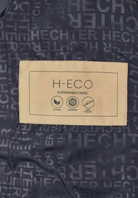 Dunkler Stoff mit einem sich wiederholenden "HECHTER"-Muster, versehen mit einem beigefarbenen Etikett, das "H-ECO" angibt, sowie Piktogrammen für Nachhaltigkeit, Umweltfreundlichkeit und Wasserabweisung.