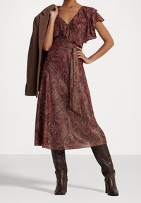 Rotes Paisley-Kleid mit Rüschensäumen, gebundenem Taillenbündchen und einem schwingenden Rock, kombiniert mit einem braunen strukturierten Mantel und knielangen braunen Stiefeln.