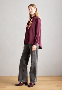 Woman standing in burgundy blouse with patterned scarf, wide gray jeans, and burgundy heeled loafers on wooden floor.