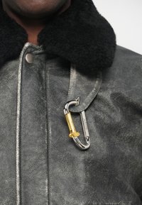 Veste en cuir texturé gris avec un col en shearling noir. Elle dispose d'un clip en carabine argenté avec un accent doré attaché à la sangle.