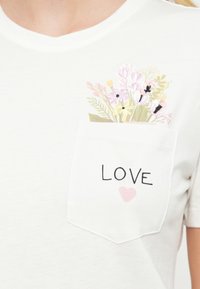 Λευκό μπλουζάκι με V-λαιμόκοψη, διακοσμημένο με κεντητό λουλουδένιο σχέδιο στην τσέπη και τη λέξη "LOVE" με λεπτομέρεια ροζ καρδιάς.