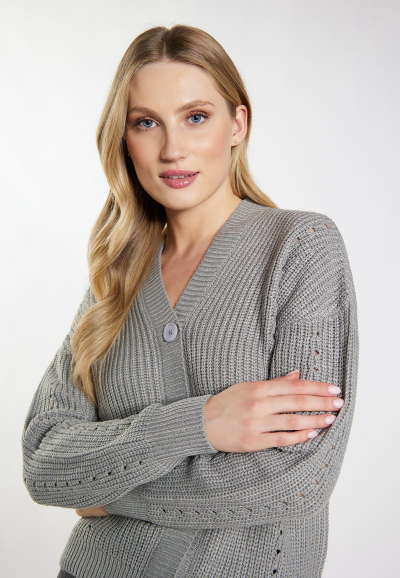 Cardigan de punto gris con escote en V, cierre de un solo botón, patrón texturizado y detalles abiertos en las mangas. Material suave con acentos acanalados.