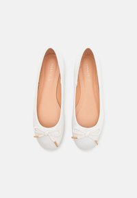 White canvas ballet flats with round toes, a decorative bow at the front, and beige insoles. Gold-tone accents on the bow detail.