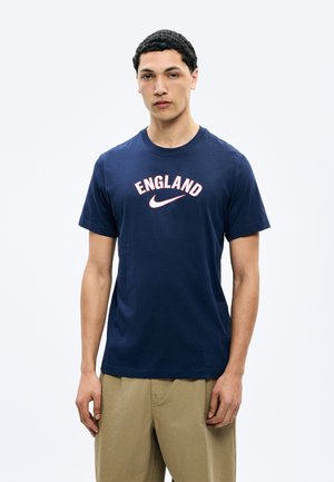 Jeune homme portant un t-shirt bleu marine avec l'inscription "ENGLAND" et un logo Nike blanc, associé à un pantalon beige, debout devant un fond uni.
