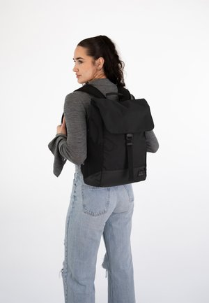 Mochila de tela negra con un diseño de rollo superior, correas ajustables y un broche frontal. Cuenta con bolsillos laterales y una textura suave.