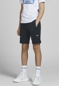 Marineblå shorts med glidelåslommer, laget av mykt materiale. Kombinert med en hvit t-skjorte med blå grafikk, og hvite joggesko med svarte detaljer.