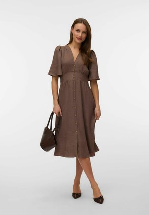 Femme portant une robe midi marron boutonnée avec des manches bouffantes, tenant un sac à main marron foncé et portant des ballerines pointues assorties.