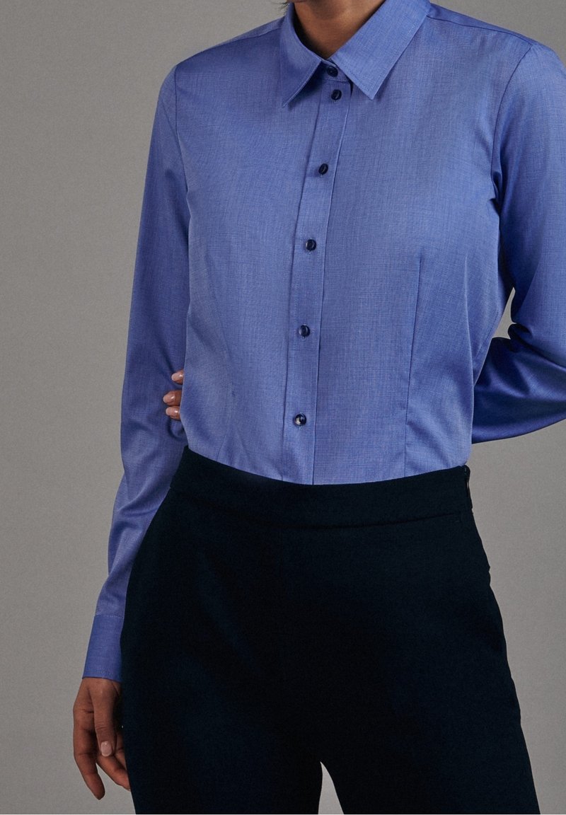 Light blue long-sleeve shirt with a collar, featuring a textured weave, button front, and a single chest pocket. Paired with dark trousers.