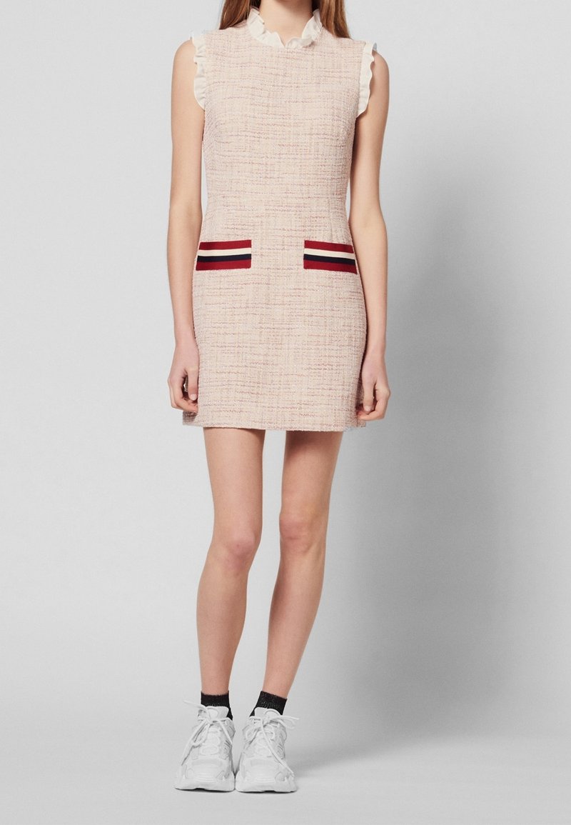 Femme portant une mini-robe sans manches rose clair texturée avec des détails de poches rayées, des chaussettes noires et des baskets blanches, debout devant un fond uni.