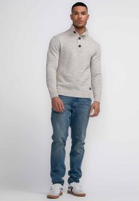 Maglione grigio chiaro a maglia con colletto alto, due bottoni neri e polsini a costine. Indossato con jeans blu e sneaker bianche con suole beige.