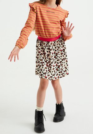 Barn iført langærmet trøje med striber og leopardmønstret nederdel, løfter en hånd, iført sorte støvler og hvide sokker, stående på hvid baggrund.