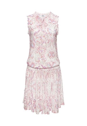 Frau mit langen Haaren, die ein ärmelloses pink-weiß gemustertes Kleid trägt, steht draußen in der Nähe von Kissen und einer Vase unter klarem Himmel.