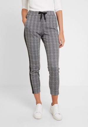 Kobieta ubrana w wąskie szare spodnie w kratę z czarnymi paskami po bokach, czarnym sznurkiem, białe trampki i biały długi rękaw.