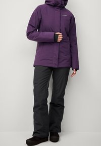 Fioletowa wodoodporna kurtka z kapturem i zapięciem z przodu, połączona z czarnymi spodniami i ciemnym obuwiem. Gładka tekstura, dopasowany krój, minimalna ilość dodatków.