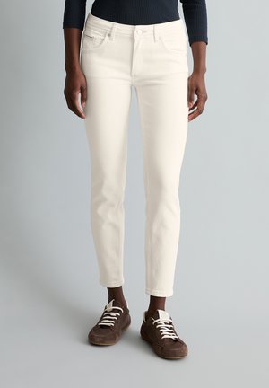 Persoon draagt off-white smalle enkelspijkerbroek en bruine vetersneakers tegen een effen lichtgrijze achtergrond.