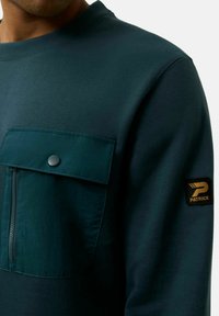 Sudadera de un tono verde oscuro con un acabado texturizado, que cuenta con un bolsillo con cremallera y un bolsillo con solapa. Incluye un parche negro de la marca en la manga.