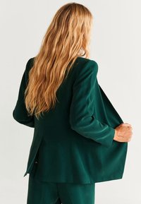 Blazer verde con un design strutturato, una texture liscia e una vestibilità su misura, indossato con pantaloni abbinati, che mette in risalto una silhouette elegante.