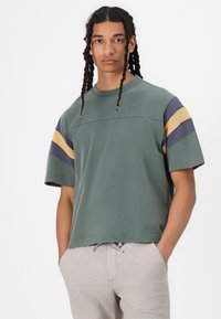 T-shirt olive à manches courtes avec des rayures horizontales bleu marine et moutarde sur les manches, col rond et coupe décontractée. Tissu en coton doux.