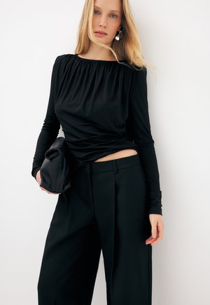 Šviesiaplaukė moteris juodu ilgarankoviu viršutiniu drabužiu ir plačiomis kelnėmis laiko sulankstytą juodą delninę, stovėdama priešais baltą foną.