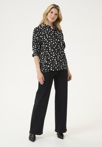 Schwarze, knöpfbare Bluse mit einem weißen abstrakten Muster, langen Ärmeln mit elastischen Bündchen, kombiniert mit schwarzen, weiten Jeans und spitzen schwarzen Schuhen.