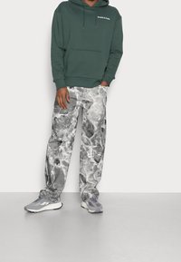 Donkergroene hoodie met een voorzak en logo, gedragen met grijs gemarmerde broek. Schoeisel zijn grijze sportieve schoenen met witte accenten.