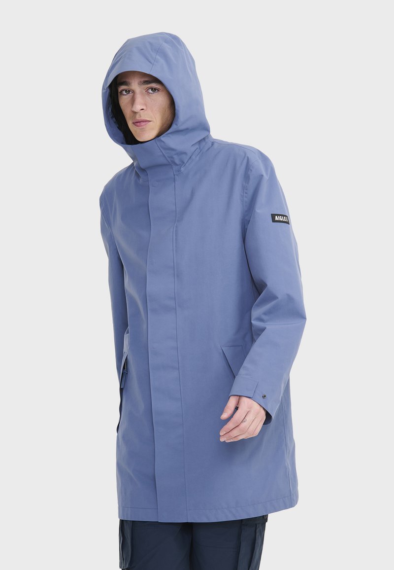 Abrigo impermeable de color azul claro con un alto cuello y capucha ajustable. Cuenta con bolsillos laterales y un parche con el logo en la manga izquierda. Textura suave.