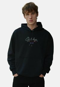 Melnā kapuce, izgatavota no mīksta auduma, ar priekšējo kabatu, kapuces auklu un krāsainiem izšuvumiem uz krūtīm.