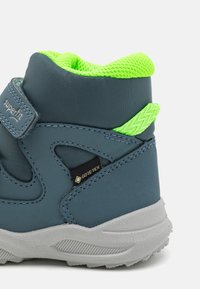 Botas infantiles impermeables de color azul con una superficie texturizada, que cuentan con un brillante color verde en el cuello de malla y cierre de Velcro. Marca Gore-Tex visible.