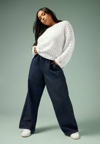 Valkoinen, pitkähihainen virkattu neule, jossa on avoin kuvio; siniset leveälahkeiset denimpantsit; valkoiset tennarit; sileä tekstuuri mintunvihreää taustaa vasten.