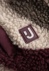 Krém, barna és bordó színű polár anyag. Texturált, bolyhos felülettel és téglalap alakú logóval rendelkezik.