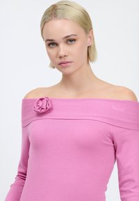 Rosa schulterfreier Pullover mit tailliertem Design, gefertigt aus einem weichen Material, mit einem Stoffblütenakzent an der Schulter.