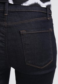 Mörkbla denimjeans med en närbild av bakfickan, som har orange sömmar och en subtil textur. Inga synliga dekorationer.