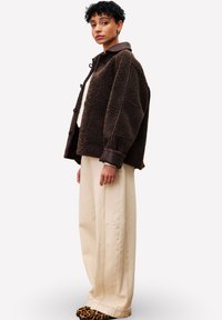 Un jeune portant une veste en polaire marron surdimensionnée, un pantalon beige large et des chaussures à imprimé léopard, se tenant devant un fond blanc uni.