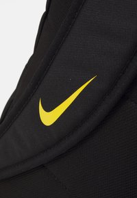 Crni ruksak od tkanine s izraženim žutim Nike logotipom, glatke teksture i strukturiranog oblika s podstavljenim rubovima.