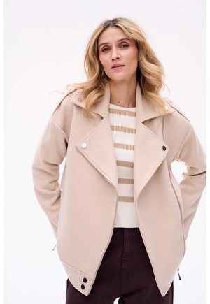 Beige Zip-Jacke mit Reversdesign, ausgestattet mit silbernen Beschlägen und Seitentaschen. Getragen über einem gestreiften cremefarbenen und beigen Pullover.