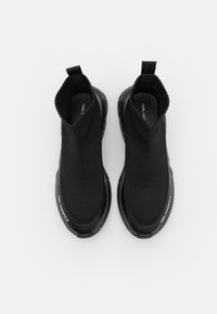 Baskets slip-on noires avec une tige en maille, texture lisse, bout arrondi et semelle amortissante. Présente un détail de logo sur le côté.