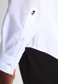 Vit långärmad skjorta med en figurnära design. Har svart rosbroderi vid manschetten och en knappstängning med en mjuk textur.