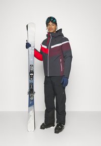 Abbigliamento da sci con una giacca a blocchi di colore nera, rossa e bianca, pantaloni neri, guanti blu e occhiali da neve, mentre tiene uno sci nero e blu.