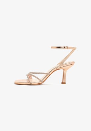 Sandalo con tacco alto a listini in oro rosa, con sottile cinturino alla caviglia e tacco medio a forma affusolata, decorato con cinturini di strass scintillanti sulle dita dei piedi.