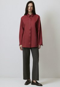 Camicia bordeaux con colletto a punta e maniche lunghe, abbinata a pantaloni grigi a gamba larga e mocassini scuri. Texture della stoffa liscia.